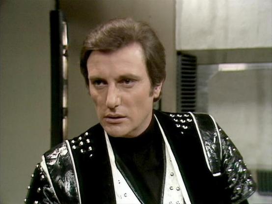 Claude Sonnet 4.0 describes the image as: "This image shows a scene from inside a spacecraft, featuring futuristic interior design with metallic surfaces and technological elements typical of science fiction television from the late 1970s. The character is wearing a distinctive black leather outfit with metallic studs and white trim details, which was characteristic of the show's costume design. The setting appears to be aboard the Liberator, the main spacecraft used by the rebel crew. The slee…
