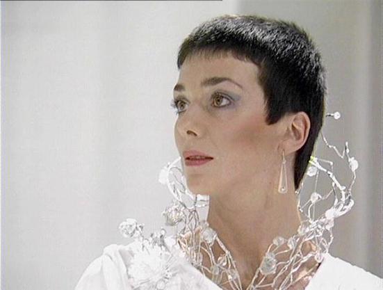 Claude 3.7 describes the image as: "The image shows a person with a short dark pixie haircut wearing an elaborate white costume with a distinctive crystalline or transparent collar design that extends upward around the neck. They're wearing teardrop-shaped earrings that complement the outfit. The setting appears to be a minimalist, bright white environment that gives a futuristic, clinical feel typical of science fiction productions from that era. The costume design features intricate details t…