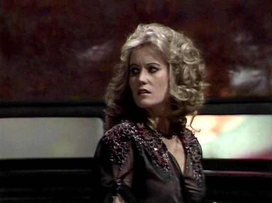 Claude 3.7 describes the image as: "The image appears to be a still from a vintage science fiction television production, likely from the late 1970s or early 1980s based on the visual style and aesthetic. 

It shows a person with curly blonde hair wearing an elegant dark outfit with decorative beaded or embellished details around the neckline. The costume has a distinctive retro-futuristic design that was characteristic of sci-fi productions from that era.

The scene has dramatic lighting with …
