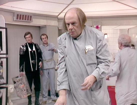 Claude 3.7 describes the image as: "This image appears to be from a science fiction television series, showing a scene set in what looks like a medical or scientific facility with white walls and ceiling. In the foreground is a character wearing a light gray uniform or lab coat who appears to be moving across the room with a stern expression. Behind him are several other characters - one in a black outfit with decorative elements, and others in gray uniforms. The setting has a clinical, futuris…
