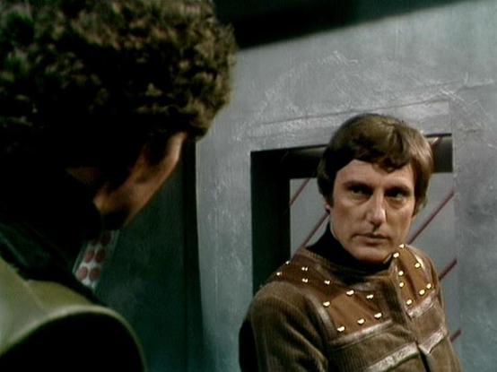 Claude Sonnet 4.0 describes the image as: "This appears to be a scene from a science fiction television series showing two characters in what looks like a spacecraft or futuristic setting. One character is wearing a green uniform or jacket and is shown from behind, while the other character faces toward the camera wearing what appears to be a brown leather outfit with metallic studs or decorative elements. The setting has the characteristic look of 1970s/80s sci-fi production design, with white…