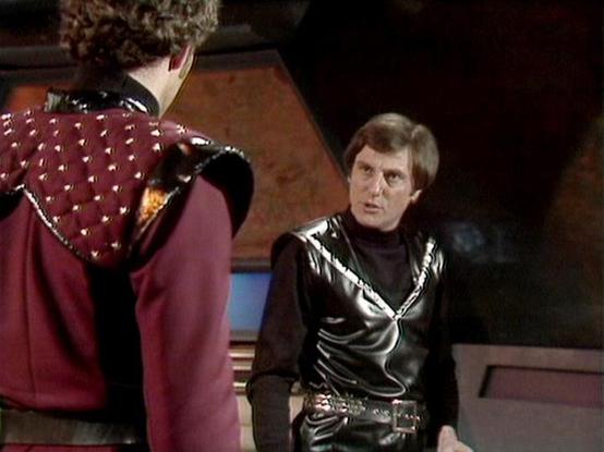 Claude 3.7 describes the image as: "The image shows a scene from a science fiction television series featuring two people in distinctive futuristic costumes. One person is wearing a burgundy outfit with studded details and is shown from behind. The other is wearing a black leather-like ensemble with silver trim and a high collar, facing the camera with a serious expression. 

The setting appears to be a spacecraft or space station interior with angular walls and technological elements visible i…