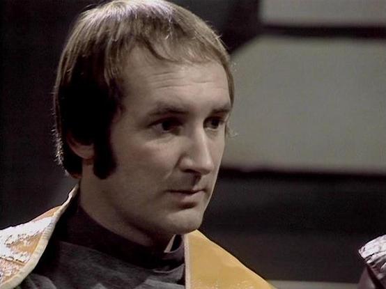 Claude Sonnet 4.0 describes the image as: "This image shows a character in what appears to be a futuristic uniform or costume, featuring dark colored fabric with distinctive yellow/gold shoulder accents or trim. The setting has a utilitarian, spaceship-like interior with curved walls and surfaces typical of science fiction television production design from this era. The lighting creates dramatic shadows and highlights that emphasize the futuristic atmosphere. The costume design suggests this is…