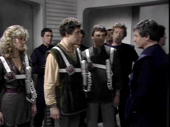 Claude Sonnet 4.0 describes the image as: "This appears to be a tense confrontation scene set in what looks like a spacecraft corridor or control room. The setting has the characteristic sterile, metallic interior design typical of space-based science fiction productions of the era. Several characters are wearing what appear to be communication devices or restraints with metallic chest pieces and coiled cables. The scene suggests a dramatic moment where characters are likely being held captive …