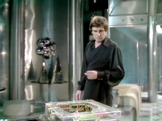 Claude 3.7 describes the image as: "This image shows a scene set in a futuristic spacecraft or space station interior with metallic walls and sci-fi styling typical of late 1970s/early 1980s British television production. 

The person in the image is wearing a dark outfit with a distinctive collar and appears to be in what looks like a control room or laboratory setting. In the foreground, there's a clear container or box with visible electronic components inside, suggesting some kind of techno…