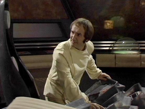 Claude 3.7 describes the image as: "This image appears to be from a science fiction television series from the late 1970s or early 1980s, based on the production style and set design. The scene takes place in what looks like the interior of a spacecraft or futuristic control room.

A person wearing a light-colored or cream jumpsuit/uniform is shown operating what appears to be some kind of technical equipment or control panel. They are inside what seems to be a retro-futuristic set with distinc…