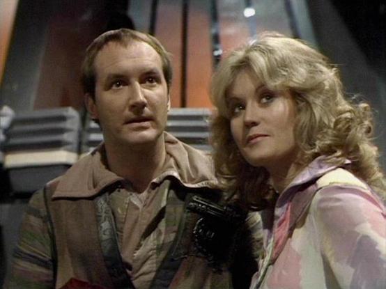 Claude Sonnet 4.0 describes the image as: "I can see two people in what appears to be a futuristic spacecraft setting, consistent with science fiction television production values of the late 1970s. The scene takes place in what looks like a ship's interior with metallic surfaces and technical equipment visible in the background. One person is wearing earth-toned clothing while the other is dressed in lighter colored attire. The lighting and composition suggest this is from a dramatic scene, wi…