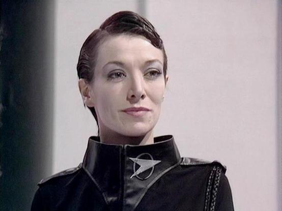 Claude 3.7 describes the image as: "This image appears to be from a science fiction television production, featuring a character in a distinctive black uniform with a high collar. The uniform includes a silver emblem or insignia on the collar that appears to be a stylized geometric shape, possibly representing some type of space organization or military faction. 

The person has short dark hair and is posed against a plain, light-colored background that gives the scene a stark, minimalist aesth…