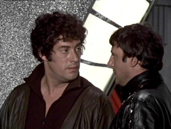 Claude 3.7 describes the image as: "This image appears to be from a classic science fiction television series from the late 1970s or early 1980s. The scene shows two individuals in what looks like a futuristic or industrial setting with a textured wall behind them. 

One person is wearing a dark brown outfit with an open collar, while the other is dressed in a black leather jacket. They appear to be engaged in a tense conversation or confrontation, facing each other closely. The lighting and fi…