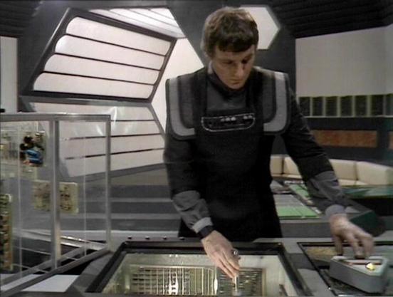 Claude 3.7 describes the image as: "This image shows a scene from a science fiction television series, featuring a person in a dark uniform with padded shoulders working at what appears to be a control console or technical station. The individual is operating some kind of electronic equipment in what looks like a spacecraft or futuristic control room.

The set design has the distinctive aesthetic of late 1970s/early 1980s British sci-fi, with angular white walls, geometric patterns, and a clean…