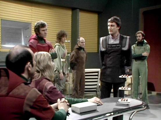 Claude Sonnet 4.0 describes the image as: "This appears to be a scene set in what looks like a spaceship or futuristic facility interior. The setting features typical sci-fi television production design with metallic walls, control panels, and utilitarian furnishing. Several characters are gathered in what appears to be a meeting or briefing scene. The costumes are characteristic of late 1970s/early 1980s science fiction television, with some characters wearing practical military-style clothing…