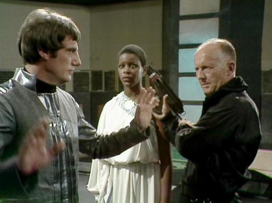 Claude Sonnet 4.0 describes the image as: "This scene appears to be set in a futuristic interior, likely aboard a spacecraft or space station, with characteristic sterile walls and technological elements typical of science fiction television. The setting has a clinical, institutional feel with neutral-colored panels and structured lighting.

The scene depicts what appears to be a tense confrontation or negotiation between three characters. One figure is dressed in dark leather-like clothing sug…