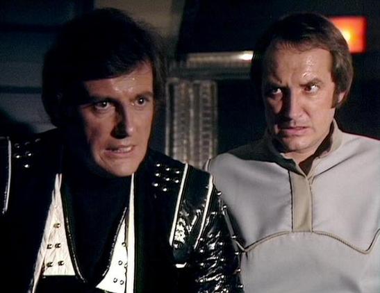 Claude 3.7 describes the image as: "This image appears to be from a science fiction television series, showing two people in a futuristic setting. On the left is a person wearing a distinctive black leather outfit with metallic studs and accents, while on the right is someone in a light-colored, possibly cream or beige costume with decorative stitching. 

The scene appears to be set in what looks like a spacecraft or control room, with technical equipment visible in the background. The lighting…
