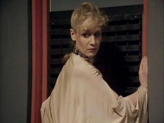 Claude 3.7 describes the image as: "The image appears to be from a vintage science fiction television production, likely from the late 1970s or early 1980s based on the filming style and aesthetic. 

It shows a person with blonde hair styled in an updo, wearing a light-colored flowing garment with wide sleeves that resembles a robe or cape. They are looking over their shoulder with a serious expression while standing near what appears to be some kind of technological equipment or console with d…