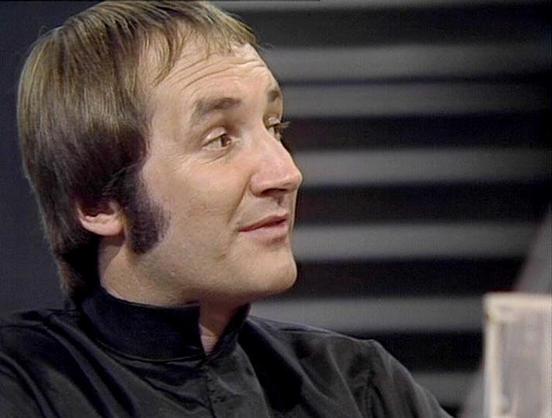 Claude 3.7 describes the image as: "The image shows a person in what appears to be a science fiction television series from the late 1970s or early 1980s. They are wearing a dark, possibly black outfit with a high collar that has a distinctive retro sci-fi style. The person has short brown hair with sideburns, characteristic of that era's fashion.

The setting appears to be some kind of spacecraft or futuristic installation with horizontal lines visible in the background. The lighting and film …