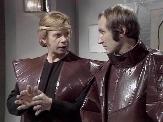 Claude 3.7 describes the image as: "The image appears to be from a science fiction television series from the late 1970s or early 1980s. It shows two people in a scene with distinctive futuristic costumes. The person on the left wears a headband and has a shiny brown or burgundy shoulder piece over a dark outfit, while gesturing with their hands during what seems to be a conversation. The person on the right is wearing a similar shiny material in what appears to be a high-collared jacket or uni…