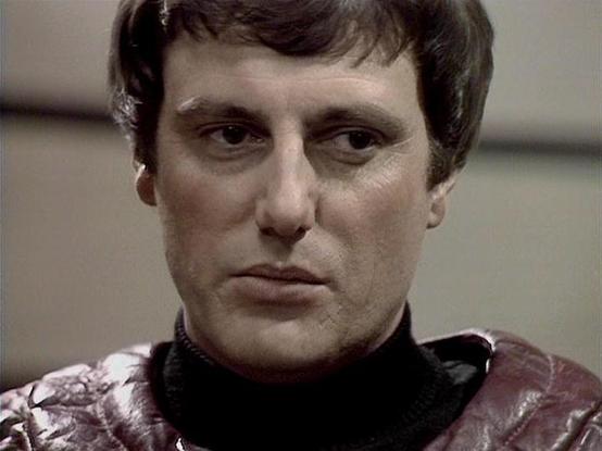 Claude Sonnet 4.0 describes the image as: "This image shows a close-up of a character in what appears to be a science fiction setting, likely aboard a spacecraft or in a futuristic facility based on the sleek, metallic background. The person is wearing distinctive costume styling typical of late 1970s/early 1980s science fiction television - a burgundy or maroon colored outfit with a high collar and what appears to be leather or vinyl material. The lighting and composition suggest this is from …