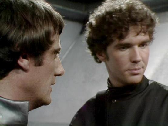 Claude 3.7 describes the image as: "This image appears to be from a science fiction television series from the late 1970s or early 1980s, showing two people in conversation in what looks like a spacecraft interior. The scene features two performers in dark clothing against a gray/metallic background, creating the futuristic aesthetic typical of sci-fi productions from that era. 

The person on the left has straight, darker hair and is shown in profile, while the person on the right has curly ha…
