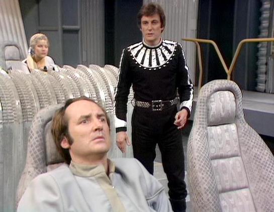 Claude 3.7 describes the image as: "This image appears to be from a science fiction television series, showing a futuristic spacecraft interior with distinctive seating. The scene features three people in a spaceship setting with padded, textured gray chairs. 

In the foreground, someone in a light-colored outfit sits in one of the chairs. Standing behind is a person wearing a striking black costume with a white decorative collar design and a belt. In the background, another figure with lighter…