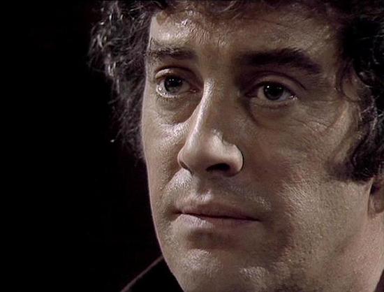 Claude 3.7 describes the image as: "This image appears to be a close-up shot from a television drama, likely from the late 1970s or early 1980s based on the filming style and visual quality. The image features a person with curly dark hair against a black background, creating a dramatic portrait with strong contrast. Their expression appears serious or contemplative, with a hint of intensity in their gaze.

The lighting technique is characteristic of sci-fi television productions from that era,…