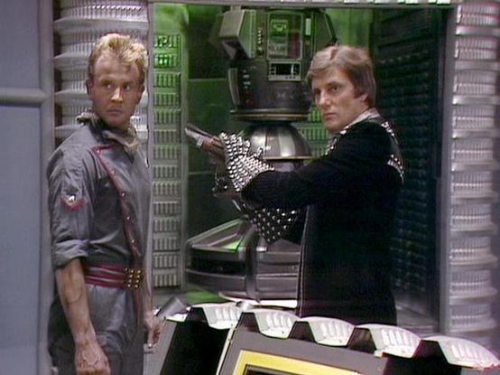 Claude 3.7 describes the image as: "This image appears to be from a science fiction television series, showing two people in what looks like a futuristic spacecraft or control room. The set features metallic walls, technical panels with green lighting, and distinctive retro sci-fi design elements typical of television productions from the late 1970s or early 1980s.

One person wears a gray uniform with rolled-up sleeves, while the other is dressed in a black outfit with studded or decorated sle…
