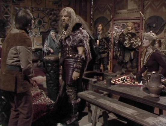 Claude 3.7 describes the image as: "This image shows a scene from a science fiction television series set in what appears to be a medieval-inspired tavern or gathering place. The setting features rustic wooden furniture, decorative elements on the walls, and a table with what looks like a game board or display.

The scene depicts several characters in distinctive costumes. In the center stands a tall figure in elaborate leather armor with fur accents and long hair. To the left is someone in sim…