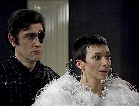 Claude 3.7 describes the image as: "This image appears to be from a vintage science fiction television series from the late 1970s or early 1980s. The scene shows two characters in distinctive costume styling. On the left is a person wearing dark clothing and an eye patch, giving them a somewhat intimidating appearance. On the right is someone with short dark hair wearing an elaborate white feathered or fluffy collar/shoulder piece with sparkly embellishments, along with large statement earrings…