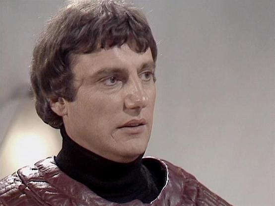 Claude Sonnet 4.0 describes the image as: "This image shows a character in what appears to be a futuristic setting, wearing a distinctive burgundy or maroon leather-style jacket with a high black collar. The costume design reflects the science fiction aesthetic typical of late 1970s British television production. The lighting and image quality suggest this is from a studio-based television production of that era. The character appears to be in a dramatic scene, with the camera capturing them in…