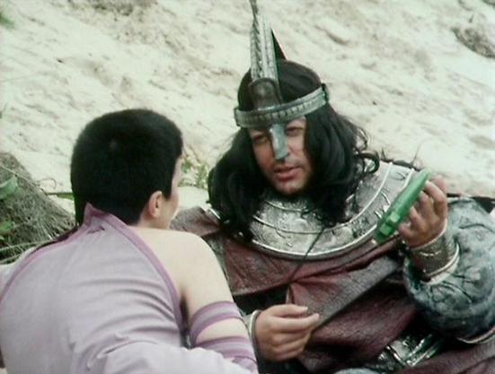 Claude 3.7 describes the image as: "The image shows a scene from what appears to be a historical or science fiction drama. One person is dressed in elaborate medieval-style armor with a distinctive spiked helmet and long dark hair. They're wearing metal plates and ornate costume elements suggesting a warrior character. This armored figure is interacting with another person in a lighter colored outfit who appears to be facing them. The scene takes place in what looks like a rocky or stone enviro…