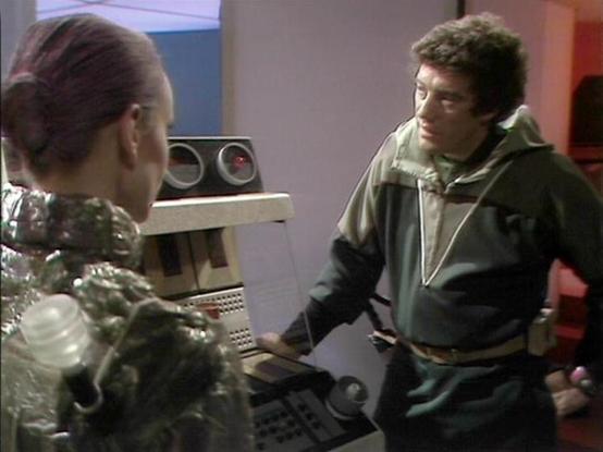 Claude Sonnet 4.0 describes the image as: "I can see this is a scene from what appears to be a science fiction television series, showing two characters in what looks like a futuristic medical or technical facility. One character is wearing metallic, reflective clothing or armor and appears to be seated at some kind of control panel or medical equipment. The other character is standing and wearing more casual, earth-toned clothing with what appears to be a hooded garment. The setting has a clin…
