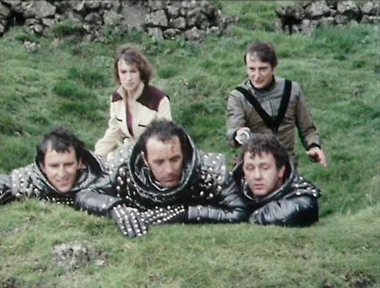 Claude Sonnet 4.0 describes the image as: "This scene appears to be set on a grassy hillside with a stone wall in the background. Three figures are lying prone on the ground wearing what looks like studded leather armor or protective gear, positioned as if they're taking cover or hiding. Two other figures are visible in the background - one wearing a burgundy and cream colored outfit, and another in lighter colored clothing. The setting has a rustic, outdoor feel typical of location shooting, w…