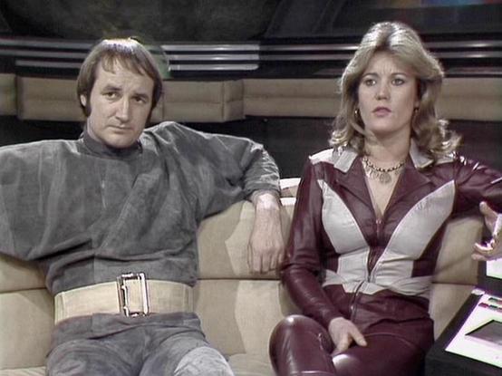 Claude Sonnet 4.0 describes the image as: "I can see two people sitting together on what appears to be a futuristic couch or seating area, likely on a spacecraft set given the technological background elements. The person on the left wears a grey/silver outfit with a distinctive belt, while the person on the right is dressed in a burgundy and white leather outfit with blonde feathered hair typical of late 1970s styling. The setting appears to be from a science fiction television production, wit…