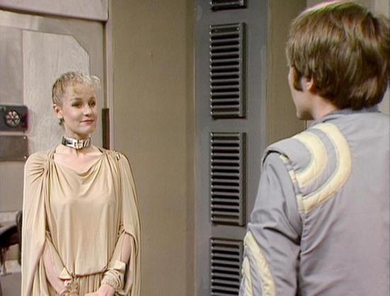 Claude 3.7 describes the image as: "This image appears to be from a science fiction television series, showing two people in a futuristic setting. On the left is a person with short blonde hair wearing an elegant cream-colored draped gown with a distinctive metallic collar. They're facing another person on the right who is partially visible from behind, wearing a gray uniform with yellow shoulder detailing.

The scene takes place in what looks like a spacecraft or space station interior, with m…