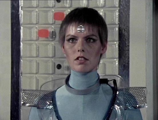Claude Sonnet 4.0 describes the image as: "I can see this is a science fiction scene featuring a person in futuristic costume and makeup. The character appears to be wearing a metallic silver and light blue outfit with distinctive shoulder pieces, and has a metallic disc or emblem on their forehead. The setting looks like the interior of a spacecraft or futuristic facility, with control panels and technological equipment visible in the background. The lighting and production design are characte…