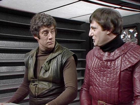 Claude Sonnet 4.0 describes the image as: "I can see two actors in what appears to be a futuristic spacecraft interior setting. The scene shows them in conversation, with one wearing earth-toned clothing and the other in a distinctive red leather outfit. The background features sleek, metallic surfaces with horizontal lines typical of science fiction set design from the late 1970s. The lighting and production values are characteristic of British television from that era. The actors appear to be…
