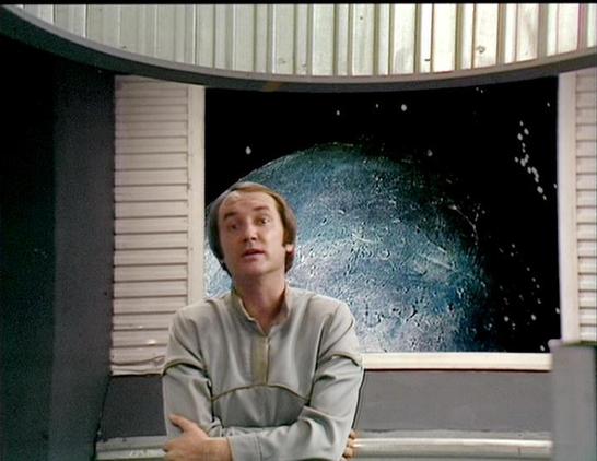 Claude 3.7 describes the image as: "This image appears to be from a science fiction television series from the late 1970s or early 1980s, based on the production style and visual effects. The scene shows a person in a light-colored tunic-style costume sitting in what looks like a spacecraft or space station interior. 

Behind them is a curved window or viewing screen displaying what appears to be a planet or moon against the darkness of space. The set design features white paneled walls typical…