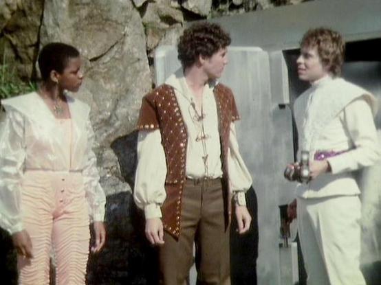 Claude 3.7 describes the image as: "This image appears to be from a vintage science fiction TV production from the late 1970s or early 1980s. The scene shows three people in costume standing together in what looks like a set with stone or rock-like features. 

The person on the left wears a cream-colored top with pink/light-colored pants. The middle figure is dressed in a brown vest with decorative elements over a white shirt with billowy sleeves and brown pants. The person on the right is wear…