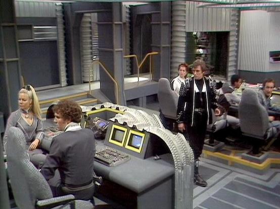 Claude Sonnet 4.0 describes the image as: "This appears to be a scene set on the flight deck or bridge of the Liberator spacecraft. The futuristic set features the characteristic curved control console with its distinctive ribbed design and multiple monitor screens. Several crew members are positioned around the bridge - some seated at the control stations while others stand nearby. The setting has the typical gray and metallic color scheme of the ship's interior, with the multi-level bridge de…