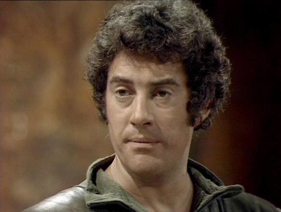 Claude 3.7 describes the image as: "The image shows a person with curly dark hair wearing what appears to be a green or olive-colored jacket with a distinctive collar. The background is blurred but suggests an indoor setting with warm, muted tones. The lighting and film quality have a distinctive vintage television production style that was common in British sci-fi programs from the late 1970s/early 1980s.

The person has a serious, contemplative expression and appears to be in a dramatic scene…