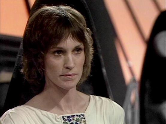 Claude 3.7 describes the image as: "The image shows a person with short brown hair wearing a white garment with a decorative pattern at the neckline. They appear to be in what looks like a futuristic or sci-fi setting, with geometric shapes and angled panels visible in the background, which has a warm orange/pink tone.

The style of the costume and set design is characteristic of British television science fiction from the late 1970s to early 1980s, with its distinctive aesthetic and production…