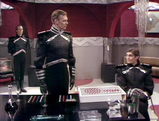 Claude 3.7 describes the image as: "This image shows a scene set in what appears to be a futuristic command center or ship interior with distinctive red and silver/gray color scheme. Three individuals are present, all wearing similar dark military-style uniforms with metallic accents and trim. The uniforms have a formal, high-ranking appearance with structured shoulders and decorative elements.

Two officers are standing while one is seated at what looks like a control console or table with col…