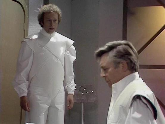 Claude Sonnet 4.0 describes the image as: "I can see two figures in what appears to be a futuristic interior setting with clean, white and beige walls typical of science fiction production design. One person is wearing a distinctive all-white outfit with puffy, padded segments and a high collar that gives it an almost ceremonial or protective appearance. The other figure is dressed in white with some dark trim details. The lighting and set design create a sterile, high-tech atmosphere character…
