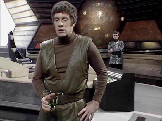 Claude 3.7 describes the image as: "This image shows a scene from a science fiction television series set on what appears to be a spacecraft or control room. The foreground shows a person wearing a brownish-olive outfit with a utility belt, standing near what looks like a control console. In the background, another figure can be seen wearing a metallic-looking jacket, standing in a more shadowed area of the set.

The futuristic set design features angled walls, technical equipment, and distinct…