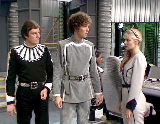 Claude 3.7 describes the image as: "This image appears to be from a science fiction television production, showing three people in distinctive futuristic costumes on what looks like a spacecraft set with metallic walls and control panels. The scene features a person on the left wearing a black outfit with a distinctive white collar design, a central figure in a light gray uniform with a belt, and a person on the right in a white/cream-colored outfit. The costumes have that characteristic 1980s …