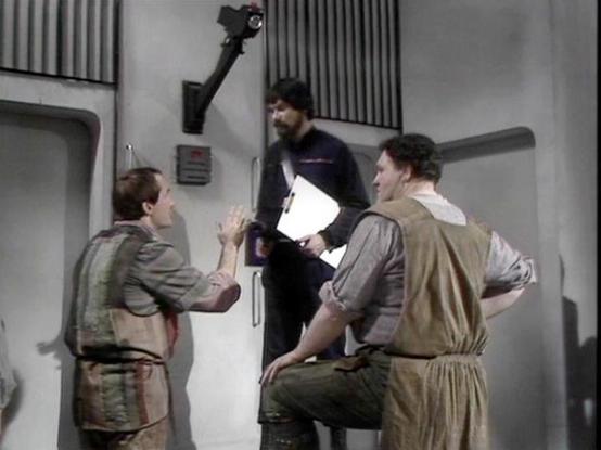 Claude Sonnet 4.0 describes the image as: "This appears to be a scene set in what looks like a spacecraft or space station corridor with characteristic futuristic wall panels and lighting. The setting has the typical industrial, utilitarian design aesthetic of science fiction television from this era. Three figures are visible in the scene - two appear to be wearing earth-toned, practical clothing that suggests they might be prisoners or rebels, while a third figure in darker clothing appears t…
