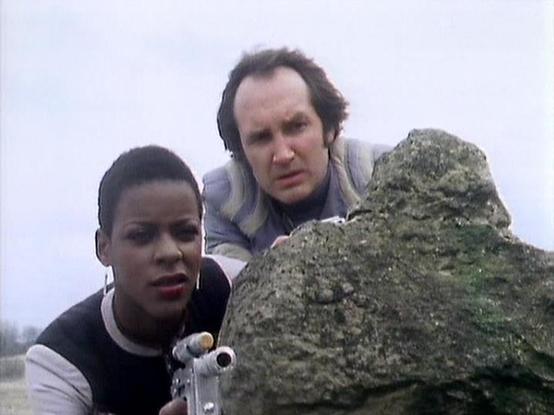 Claude Sonnet 4.0 describes the image as: "I can see two people in what appears to be an outdoor setting with rocky terrain. One person is wearing dark clothing and appears to be holding some kind of device or equipment, while another person in lighter colored clothing is positioned behind them near a large moss-covered rock formation. The setting looks rugged and natural, with an overcast sky visible in the background. The scene has the production quality typical of science fiction television …