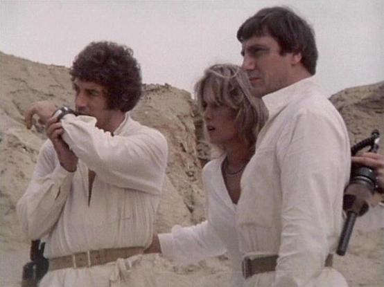 Claude 3.7 describes the image as: "This image appears to be from a science fiction television series from the late 1970s or early 1980s. The scene shows three people dressed in similar white outfits with belts in what looks like a desert or rocky terrain setting. The costumes have a distinctive retro sci-fi aesthetic typical of productions from that era.

The group appears to be in some kind of tense situation, possibly on an alien planet or remote location. One person on the left is examining…