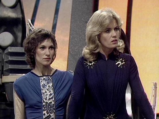 Claude Sonnet 4.0 describes the image as: "I can see this appears to be from a science fiction television series, showing two women in what looks like a futuristic setting with a distinctive sci-fi interior backdrop. One woman has shorter dark hair and is wearing a blue outfit, while the other has blonde hair styled in a 1970s fashion and is wearing a dark outfit with decorative elements. The setting appears to be inside a spacecraft or futuristic facility, with characteristic retro-futuristic …
