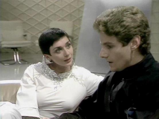 Claude 3.7 describes the image as: "This image appears to be from a vintage science fiction television production, likely from the late 1970s or early 1980s based on the filming style and aesthetic. 

The scene shows two people in conversation in what looks like a futuristic setting with textured white walls. One person wears an elegant white outfit with decorative embellishments at the neckline, while the other is dressed in a dark, possibly black outfit. 

The minimalist, clinical-looking bac…