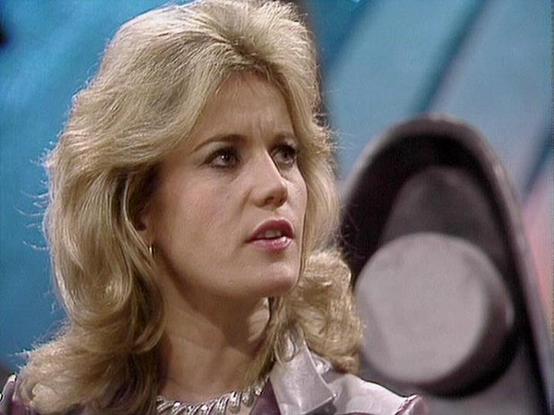 Claude Sonnet 4.0 describes the image as: "I can see this is an image from a television production, likely from the late 1970s or early 1980s based on the styling and image quality. The scene shows a blonde woman in what appears to be a metallic or shimmery outfit, with the characteristic feathered hairstyle of that era. The setting appears to be a futuristic or sci-fi environment, with blurred circular elements visible in the background that could be part of a spaceship or technological settin…